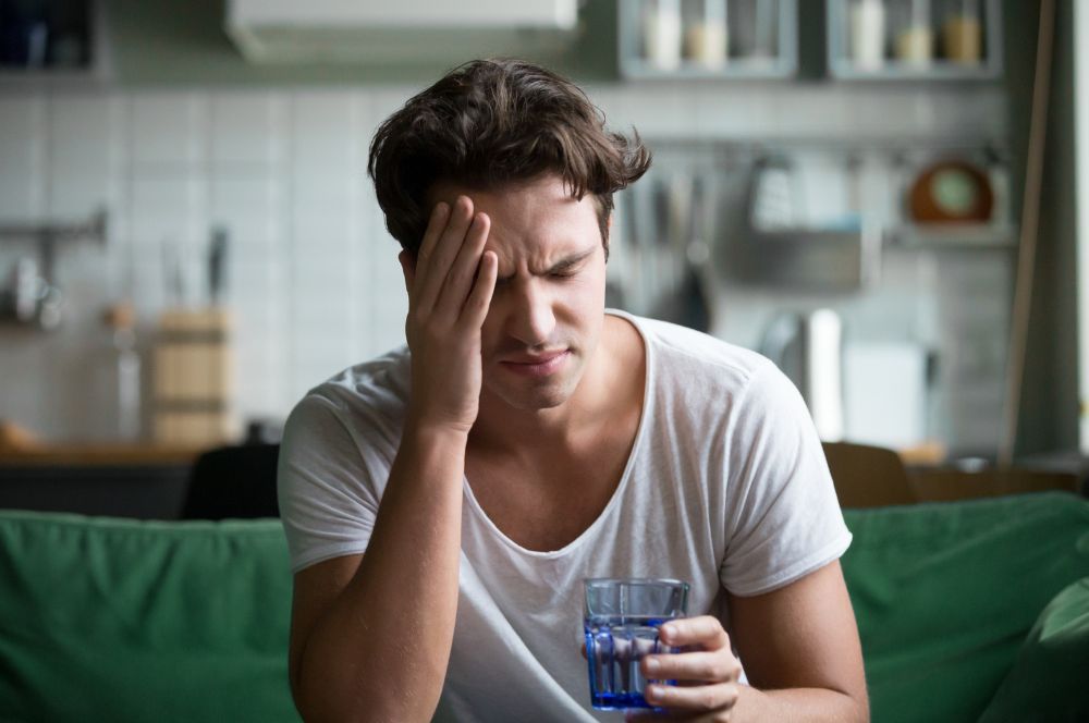 Muž sa drží za hlavu a drží v ruke pohár vody, pretože dehydratácia môže byť tiež jednou z príčin nízkeho krvného tlaku.